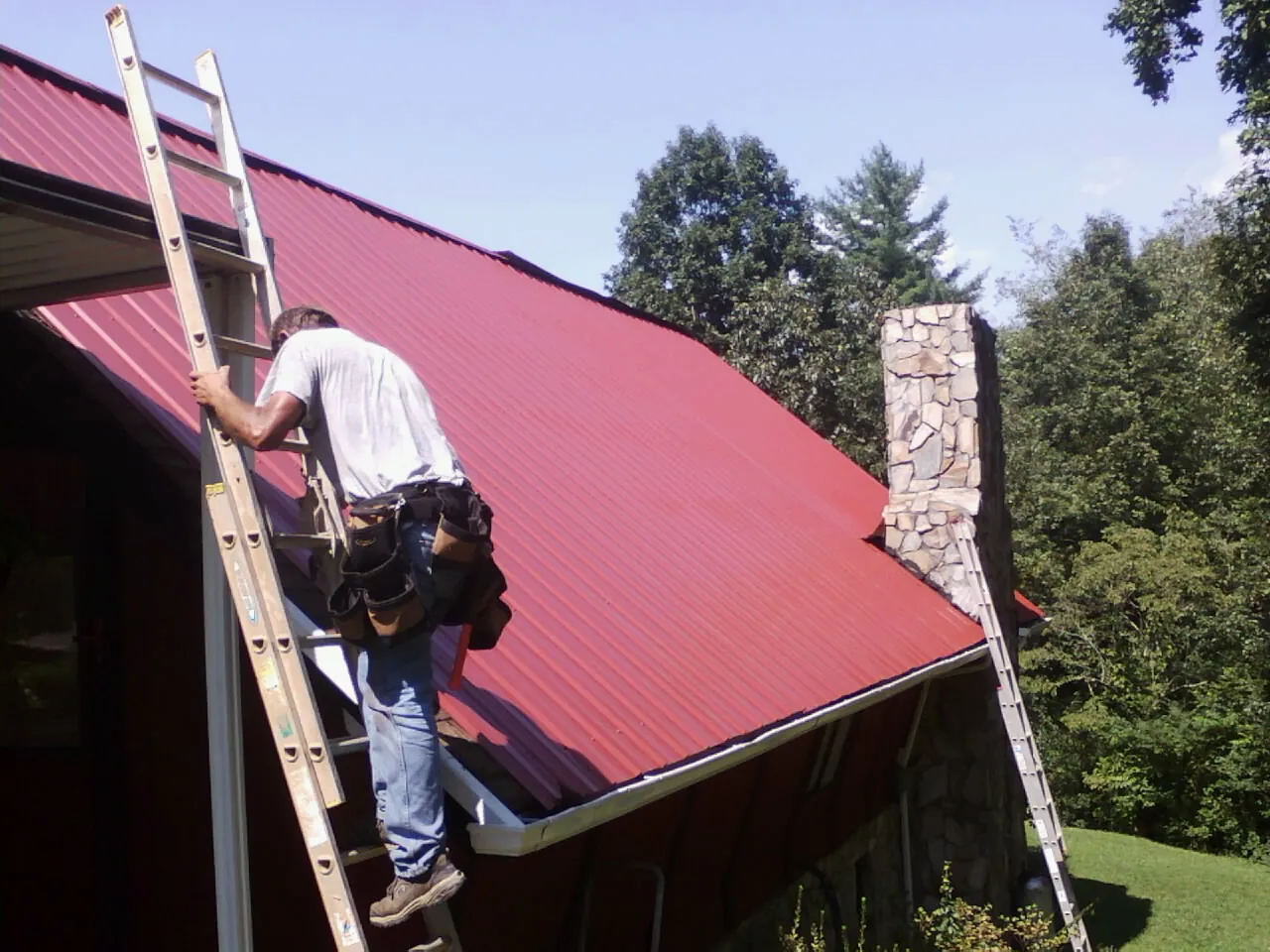Roofer on a ladder beside a red metal roof and stone chimney — Soffit & Fascia Repair by Roofing Brothers in Virginia City